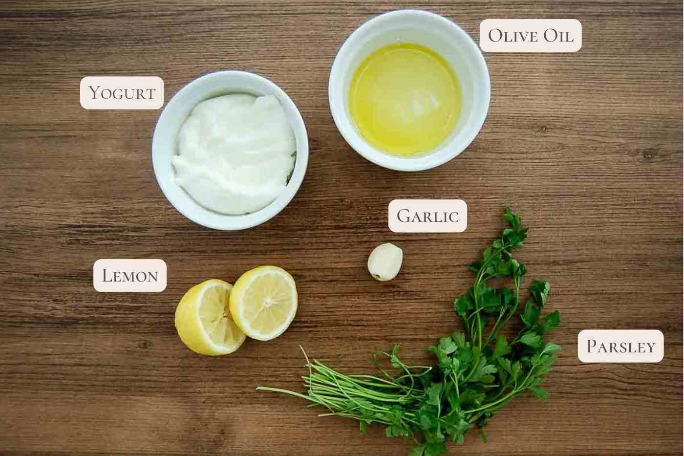 A wooden table with labeled ingredients for lemon garlic yogurt sauce: a bowl of yogurt, a bowl of olive oil, a clove of garlic, a halved lemon, and fresh parsley, all arranged neatly.