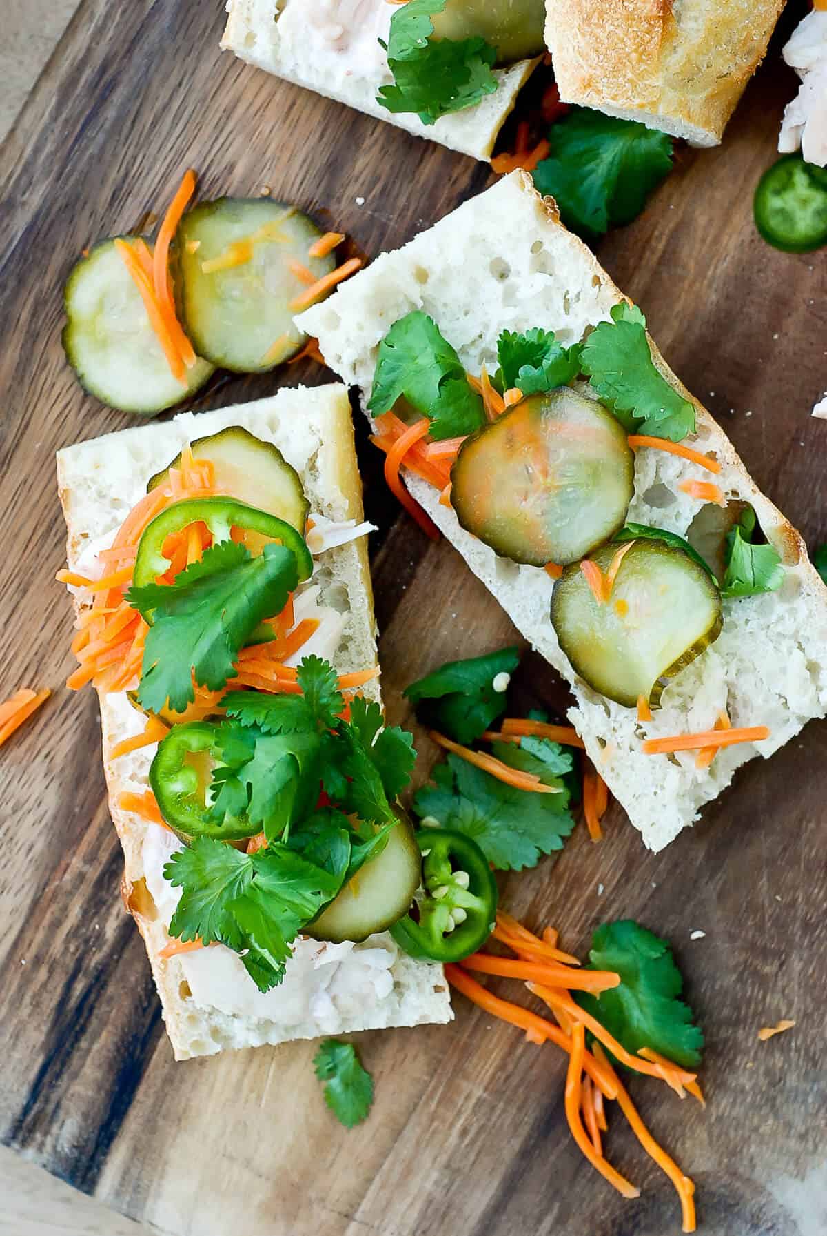 Two pieces of sliced baguette topped with classic chicken banh mi ingredients—pickled cucumbers, shredded carrots, fresh cilantro, and sliced green chili—rest on a wooden board, with scattered cilantro and carrot shreds around.