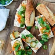 A wooden board with sliced chicken banh mi sandwiches filled with pickled cucumbers, shredded carrots, jalapeños, and cilantro sits next to a bowl of cilantro and a white napkin.