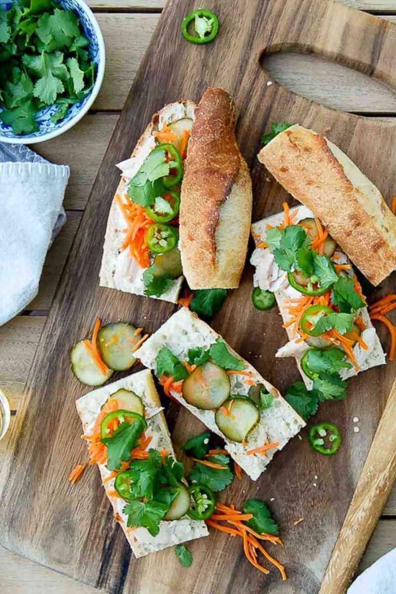 A wooden board with sliced chicken banh mi sandwiches filled with pickled cucumbers, shredded carrots, jalapeños, and cilantro sits next to a bowl of cilantro and a white napkin.