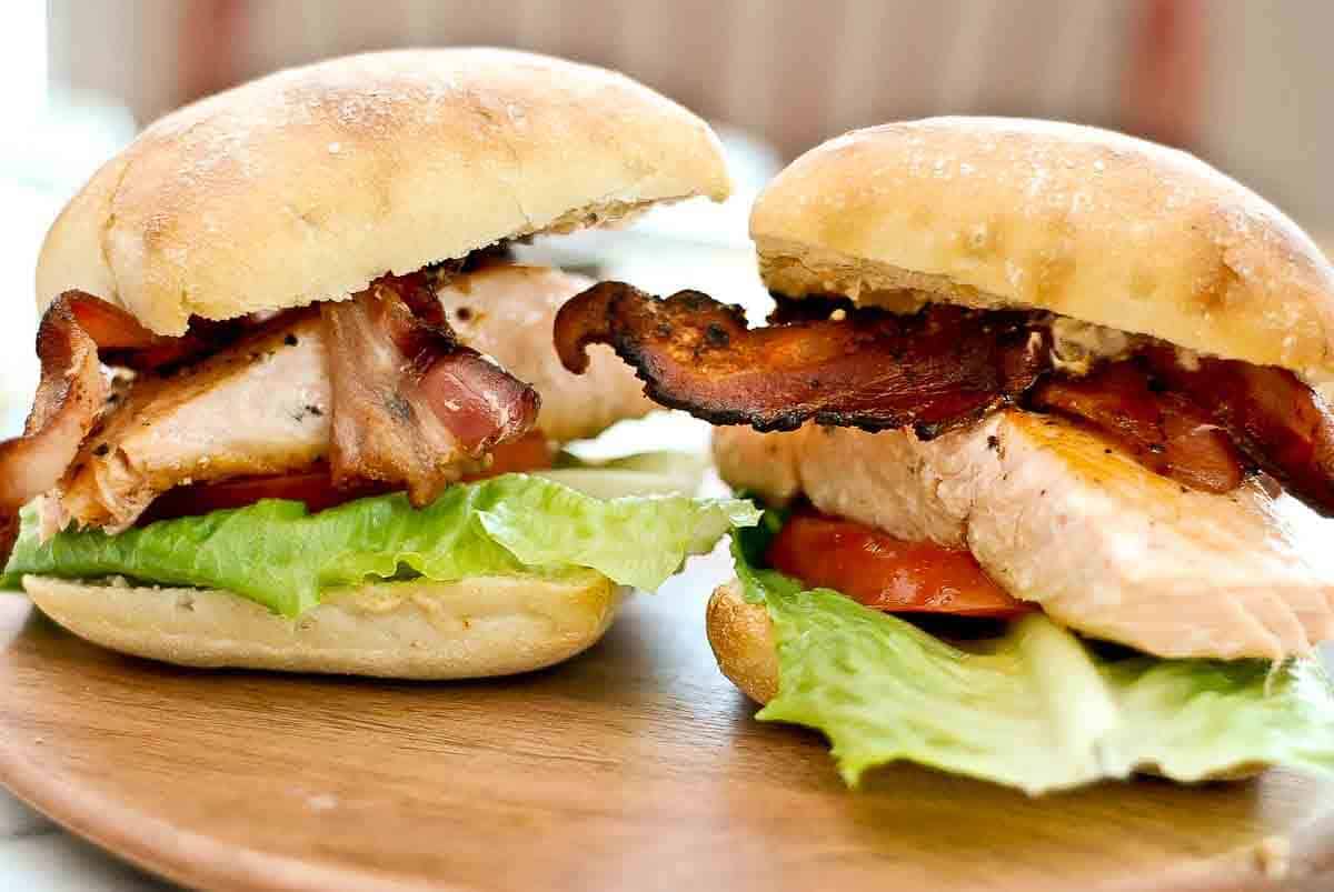 2 salmon, bacon, lettuce and tomato sandwiches on a ciabatta bun, sitting on cutting board.