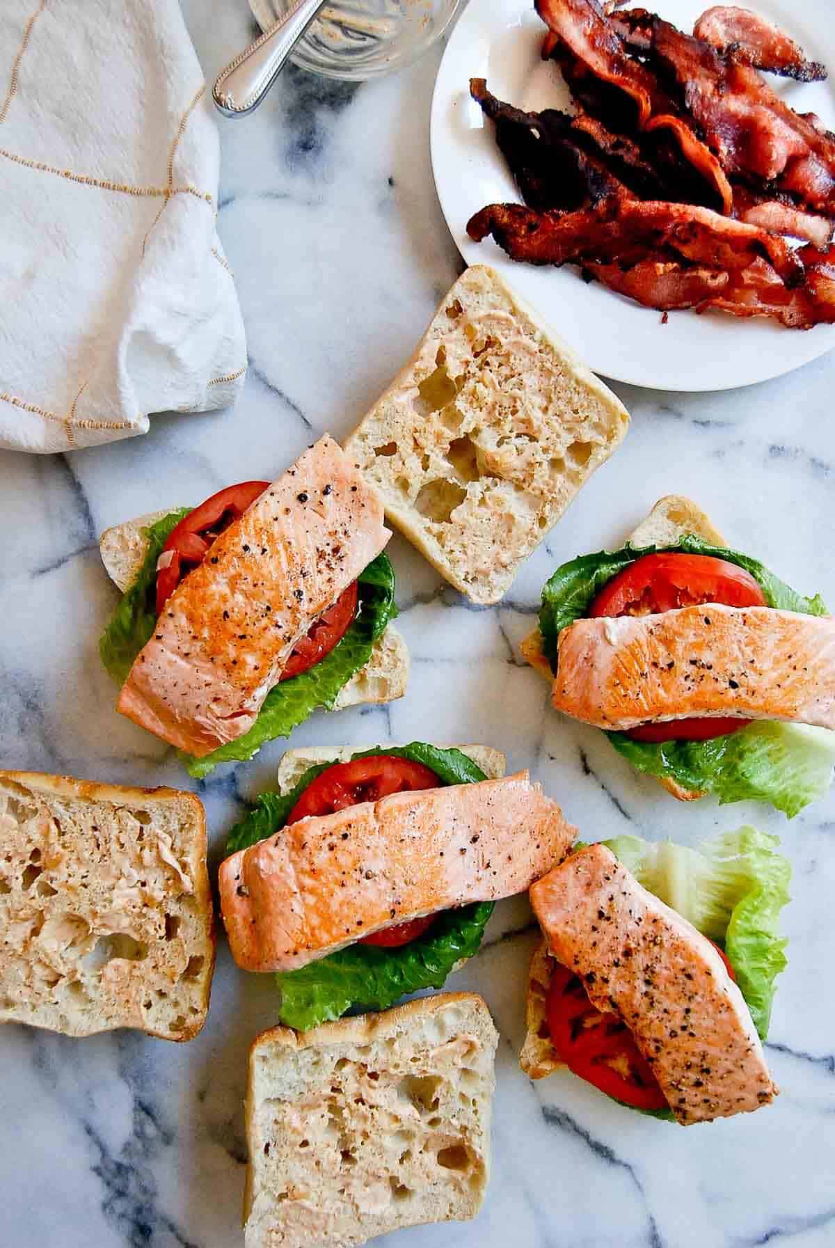 salmon pieces on buns with tomato and lettuce, and bacon on a plate to the side.