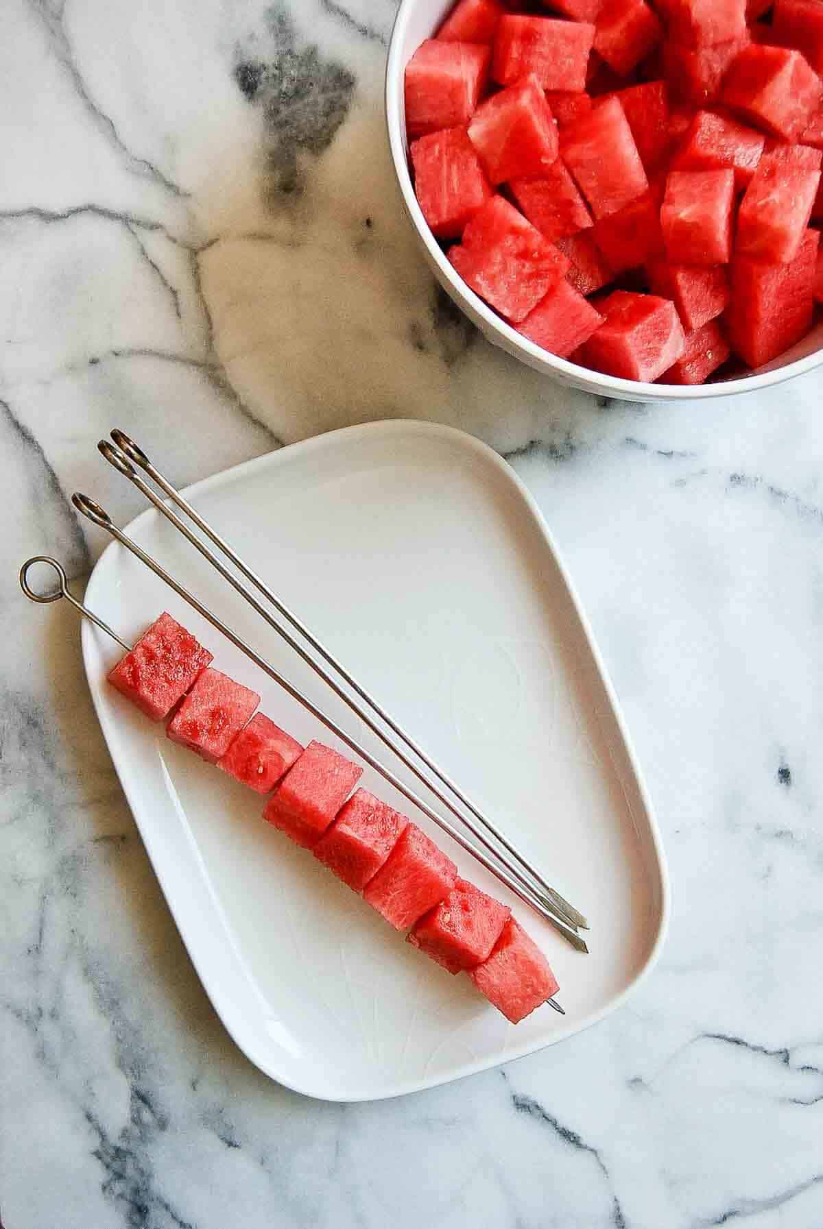 cubed watermelon on skewer with bowl of watermelon next to it.