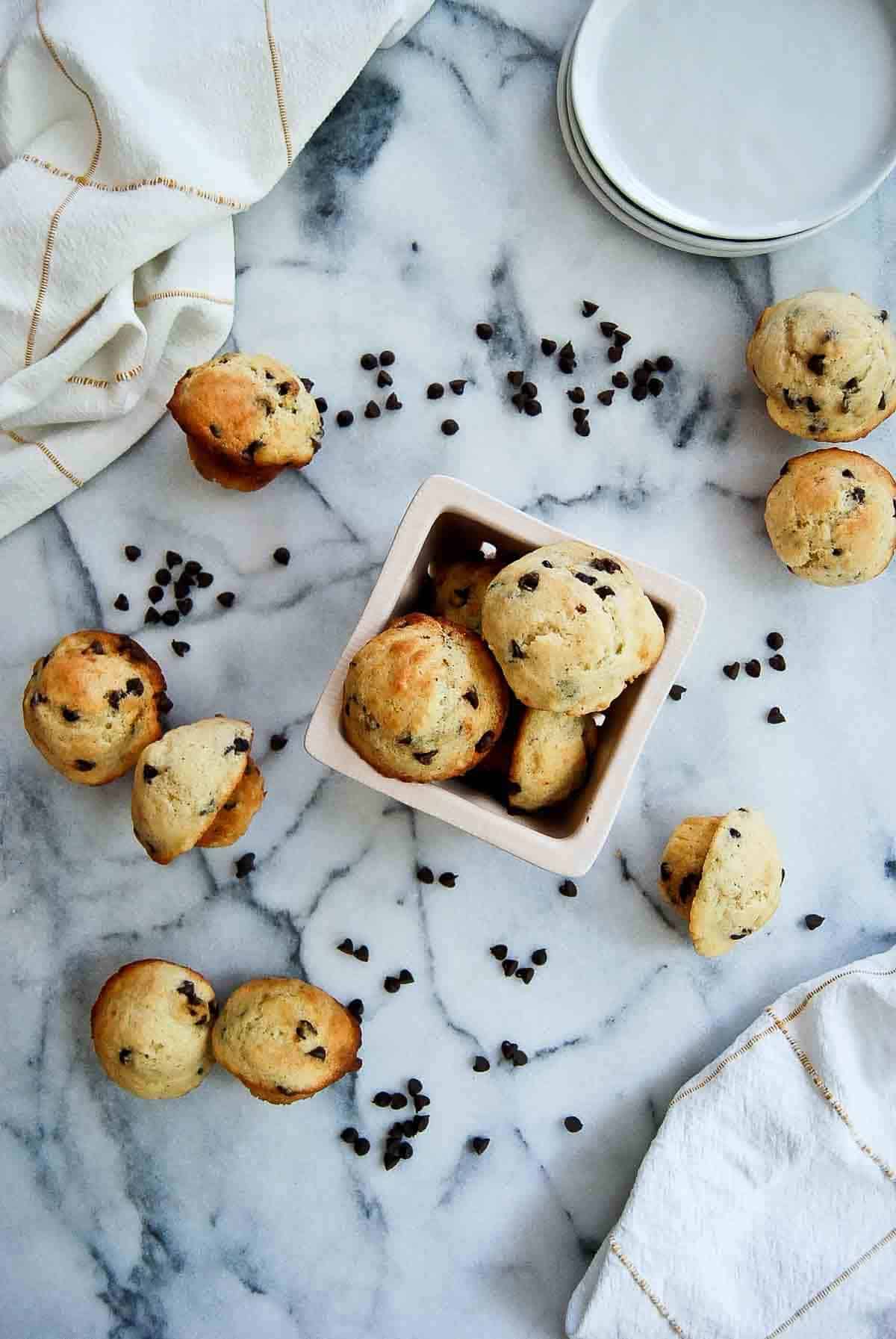 moist chocolate chip mini muffins in bowl on counter, with more muffins and chocolate chips scattered around the bowl.