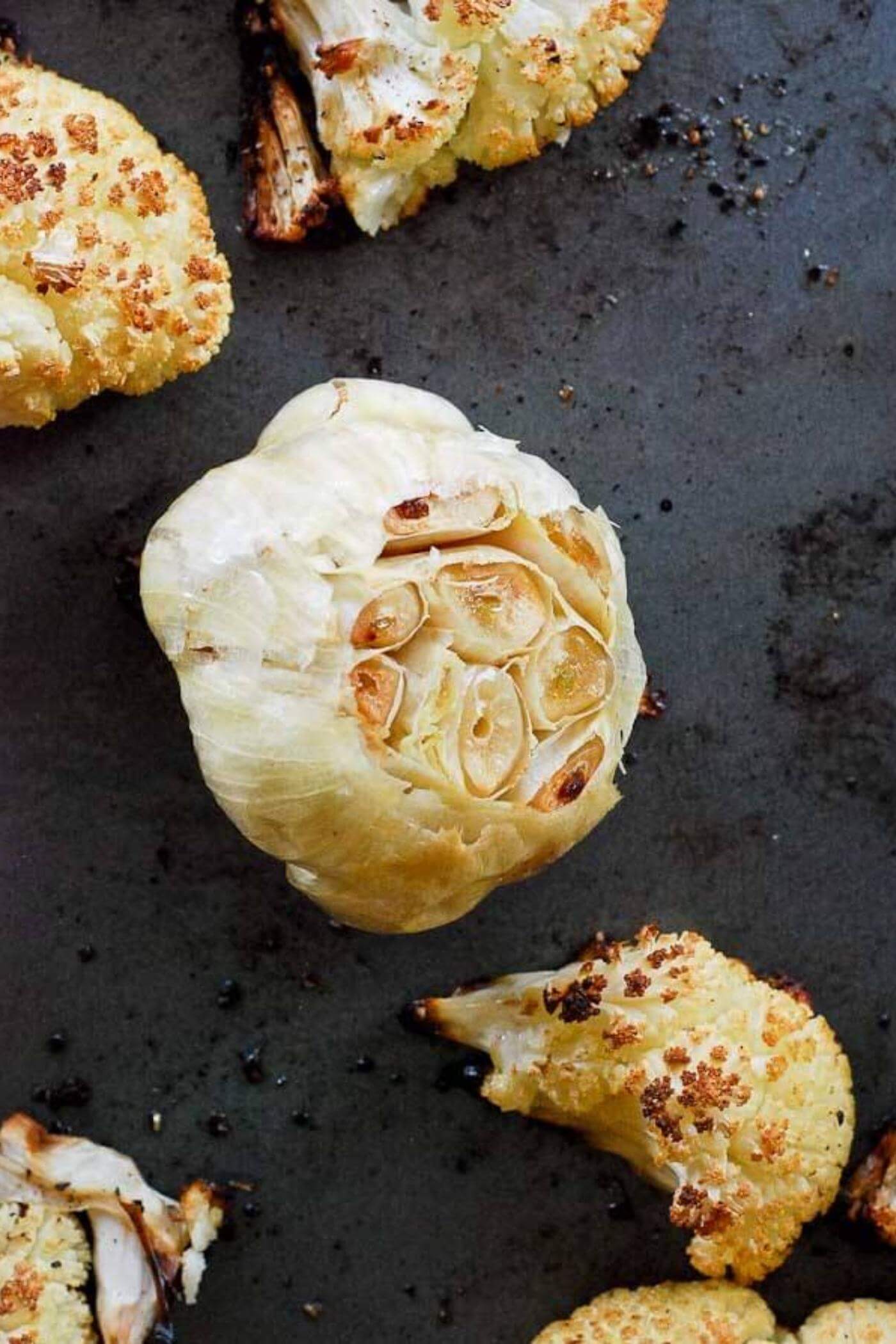 Head of roasted garlic with roasted cauliflower on baking sheet.