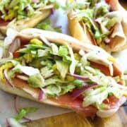 Three grinder sandwiches on a wooden board, filled with sliced deli meats, shredded lettuce, tomato, red onion, and pepperoncini—just like a classic grinder sandwich recipe, with toppings spilling out onto the board.