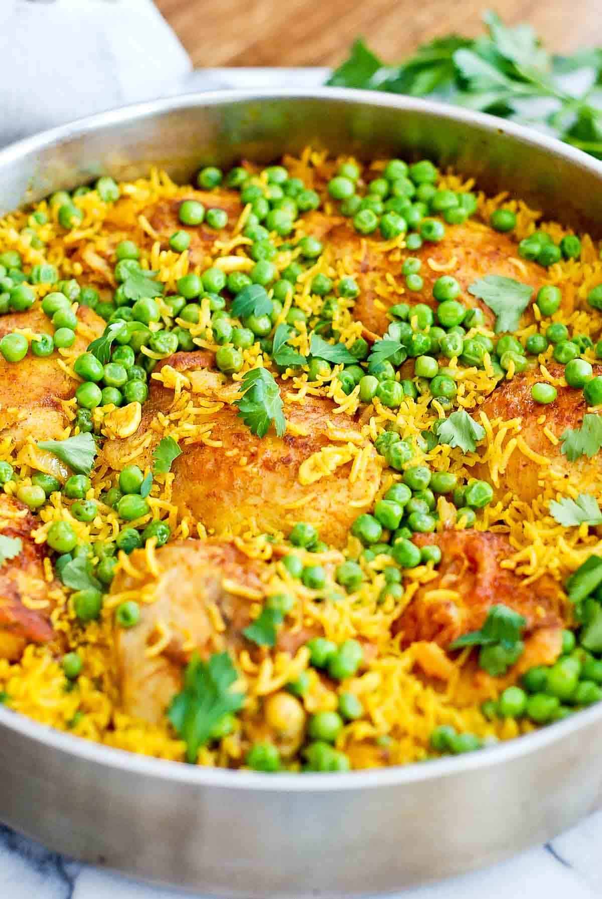 chicken and yellow rice with peas in a pan on a table with parsley in the background.