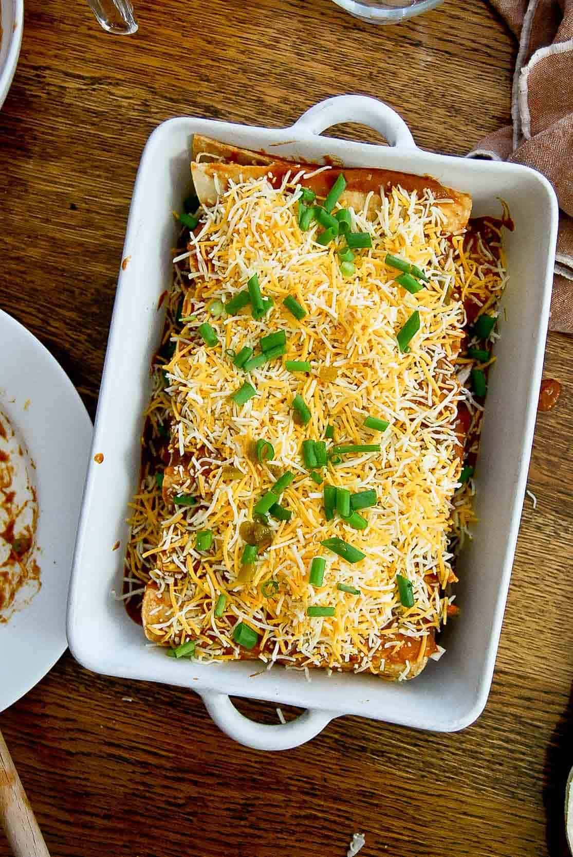 red chicken enchiladas in baking dish with shredded cheese and scallions.