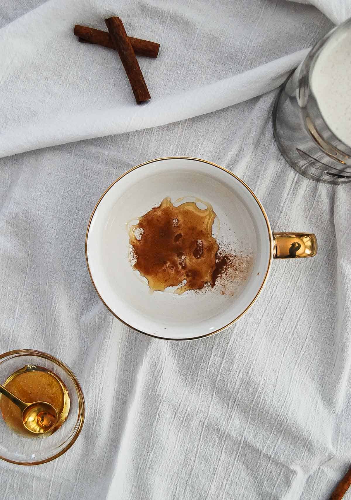 honey, cinnamon and sugar in bottom of mug on table, next to jar of honey and steamed milk.