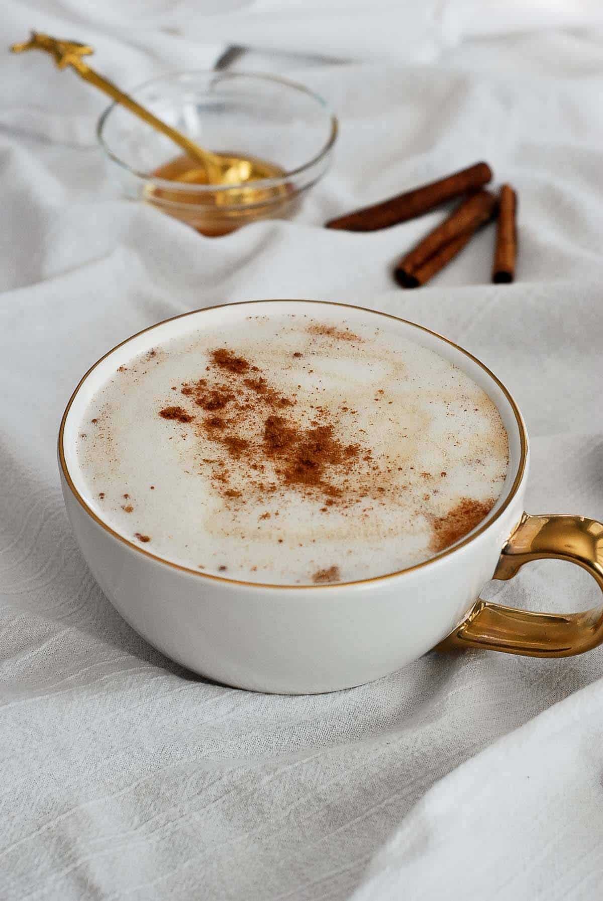 honey latte in mug with bowl of honey and cinnamon sticks in the background.