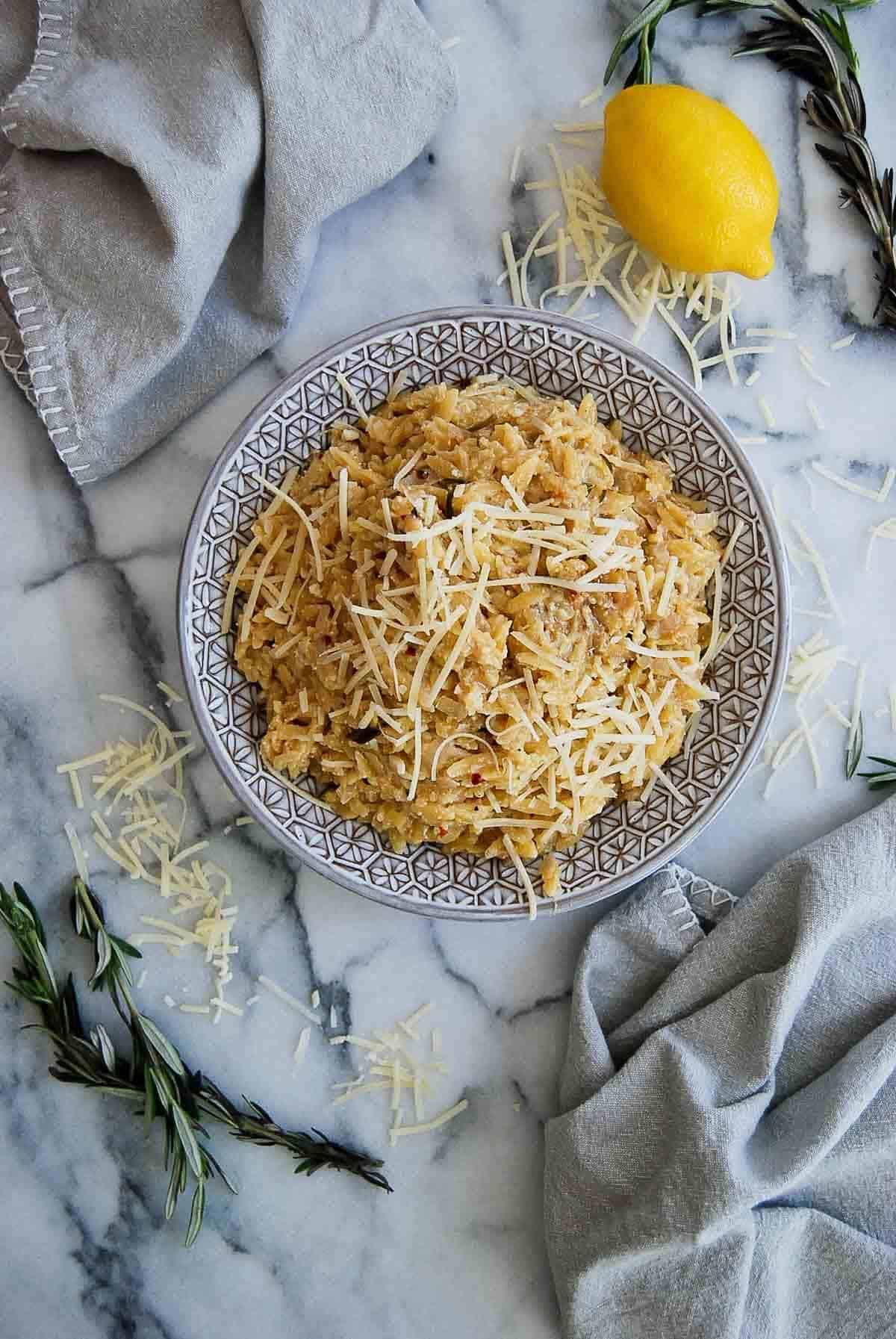 lemony orzo in serving bowl sprinkled with parmesan cheese. lemon and rosemary on the side.