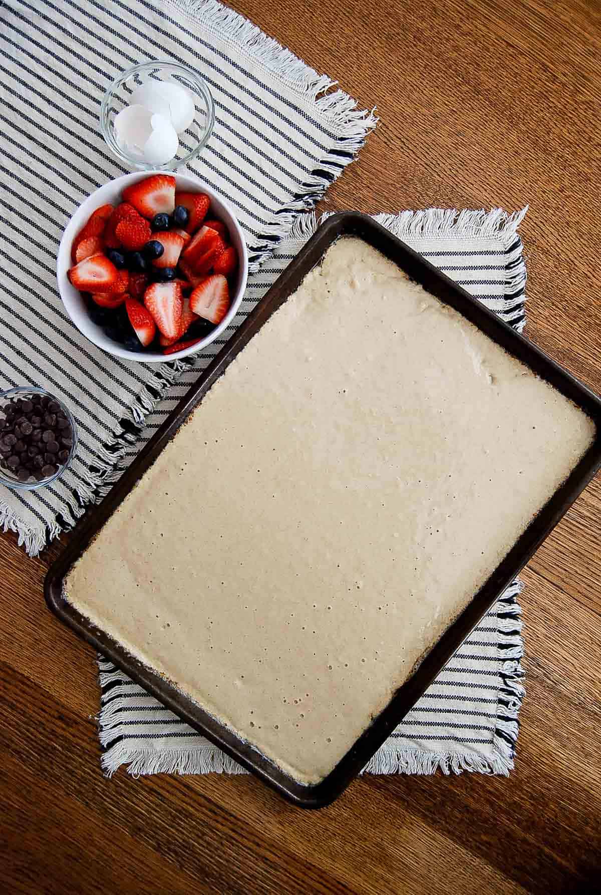 pancake batter spread evenly in baking sheet with strawberries, blueberries and chocolate chips in bowls to the side.