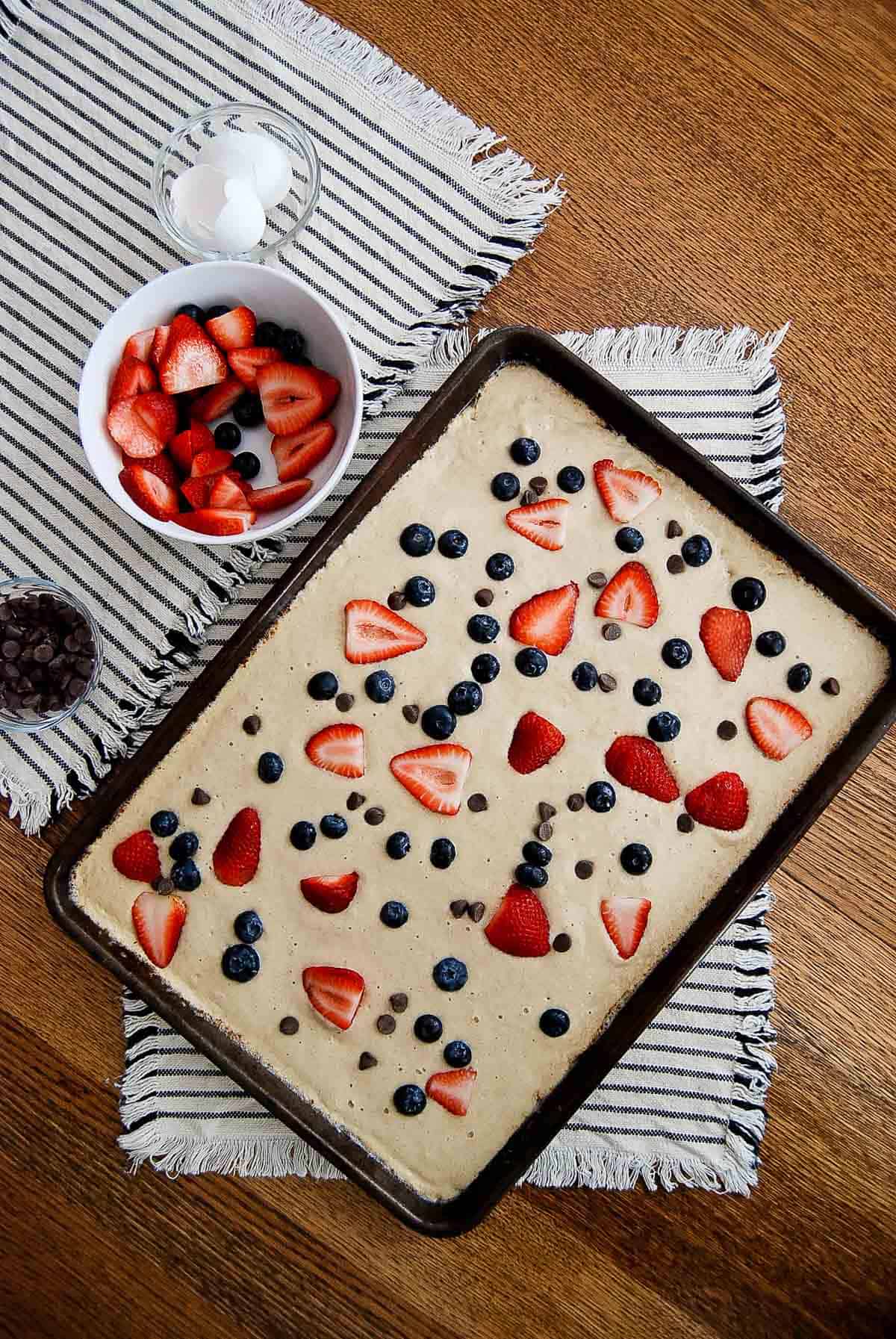 pancake batter spread in sheet pan topped with strawberries, blueberries and chocolate chips.