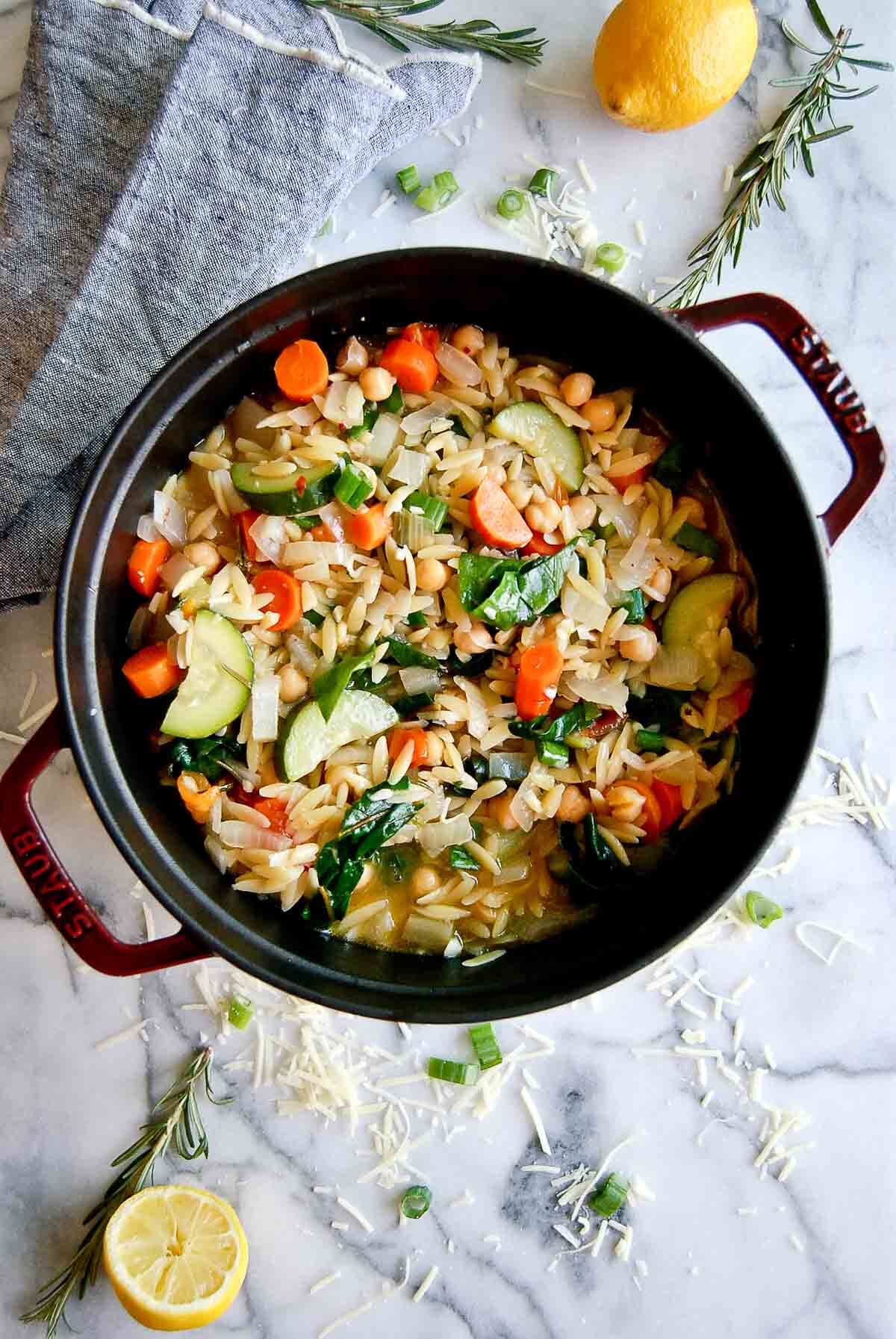 chickpea stew with orzo, veggies and greens in pot with lemons and rosemary on the sides.