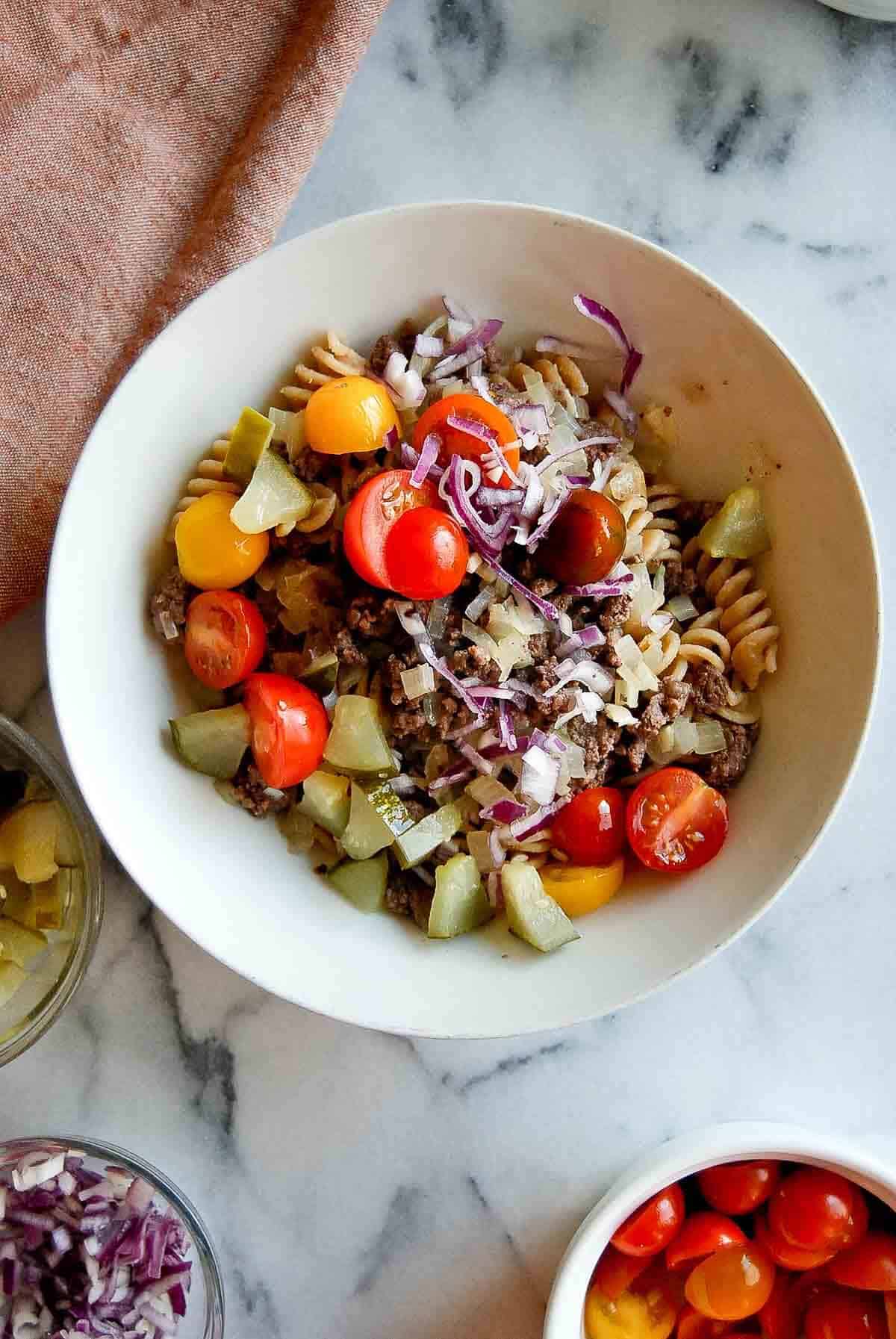 Whole wheat pasta salad with ground beef, tomatoes, onions and pickles in bowl.