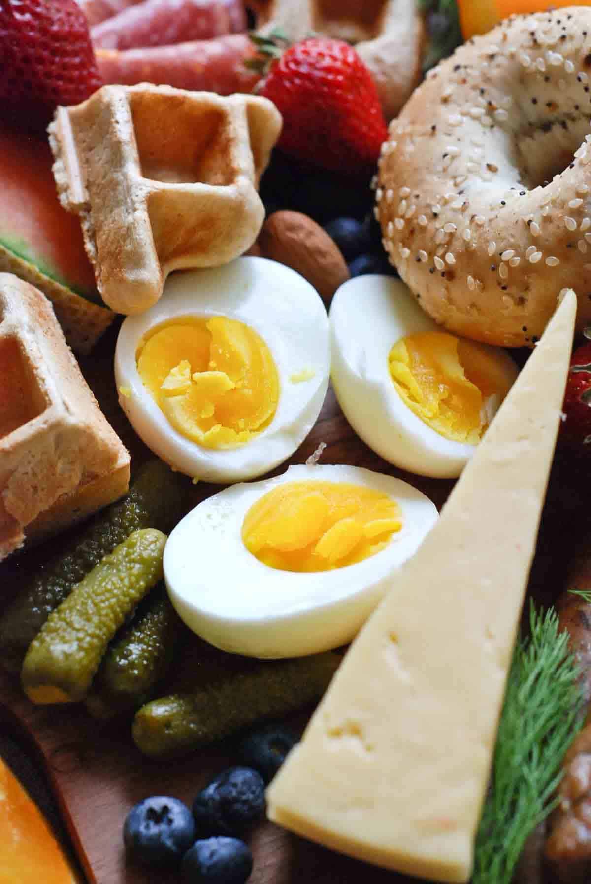 close up of jammy eggs, waffles, bagels, cheese, berries and cornichons.