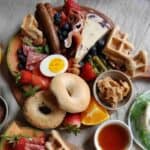 breakfast charcuterie board with egg, bagels, cheese, meat, sausage, waffles, fruit, peanut butter and syrup.