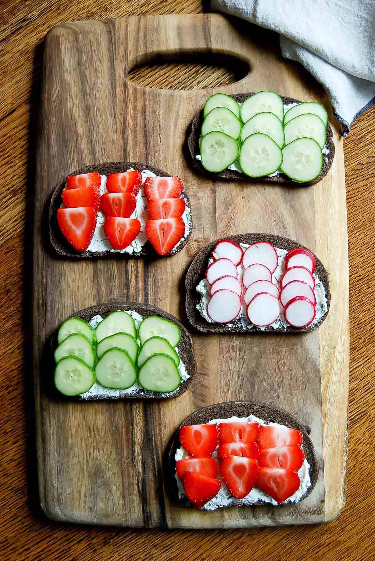 pumpernickel with herbed cheese, cucumber, strawberries and radishes on cutting board.
