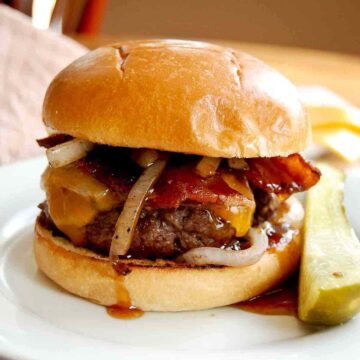 closeup view of sweet and spicy bacon burger on plate with pickle next to it.