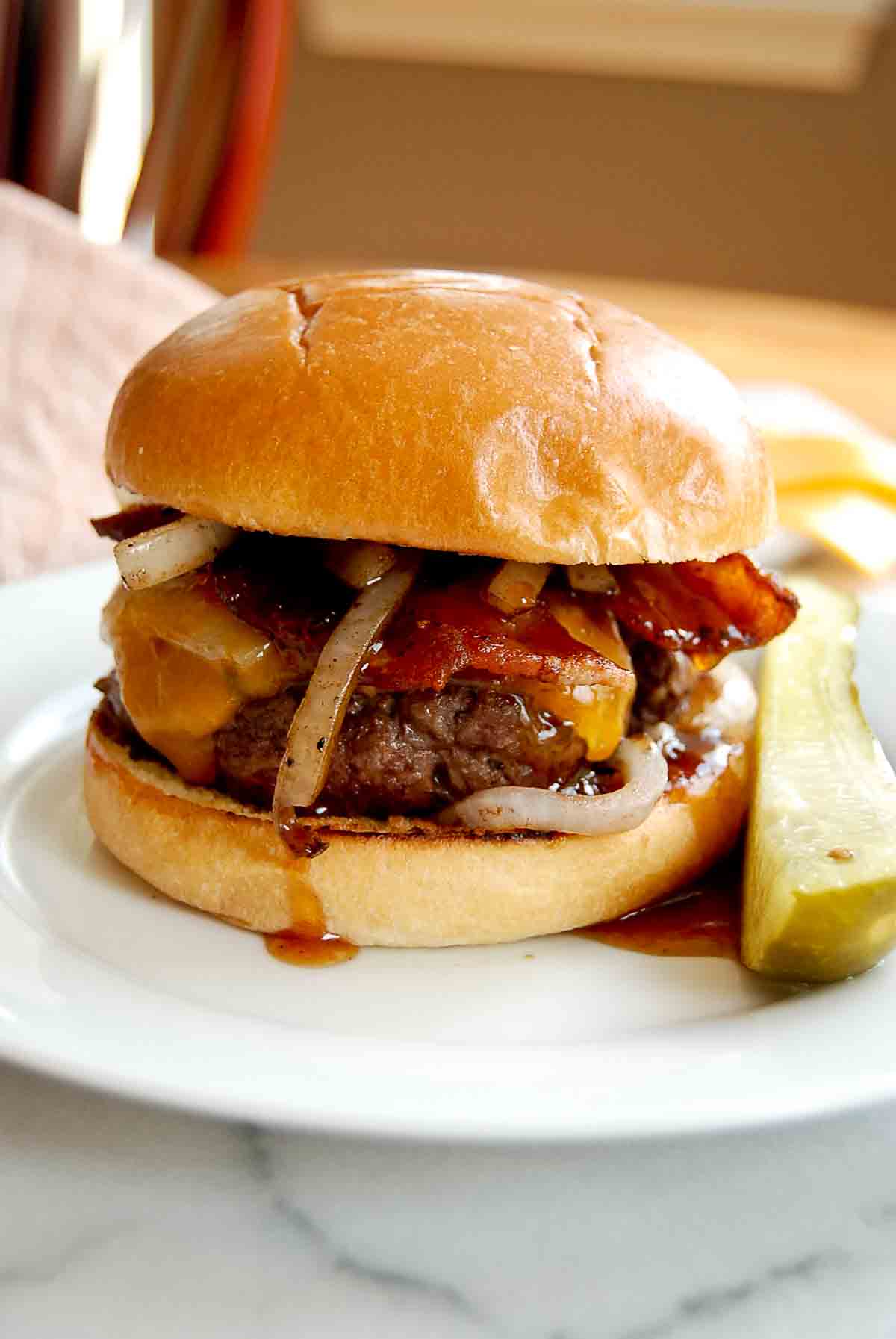 Closeup view of sweet and spicy bacon smash burger on plate with pickle next to it.