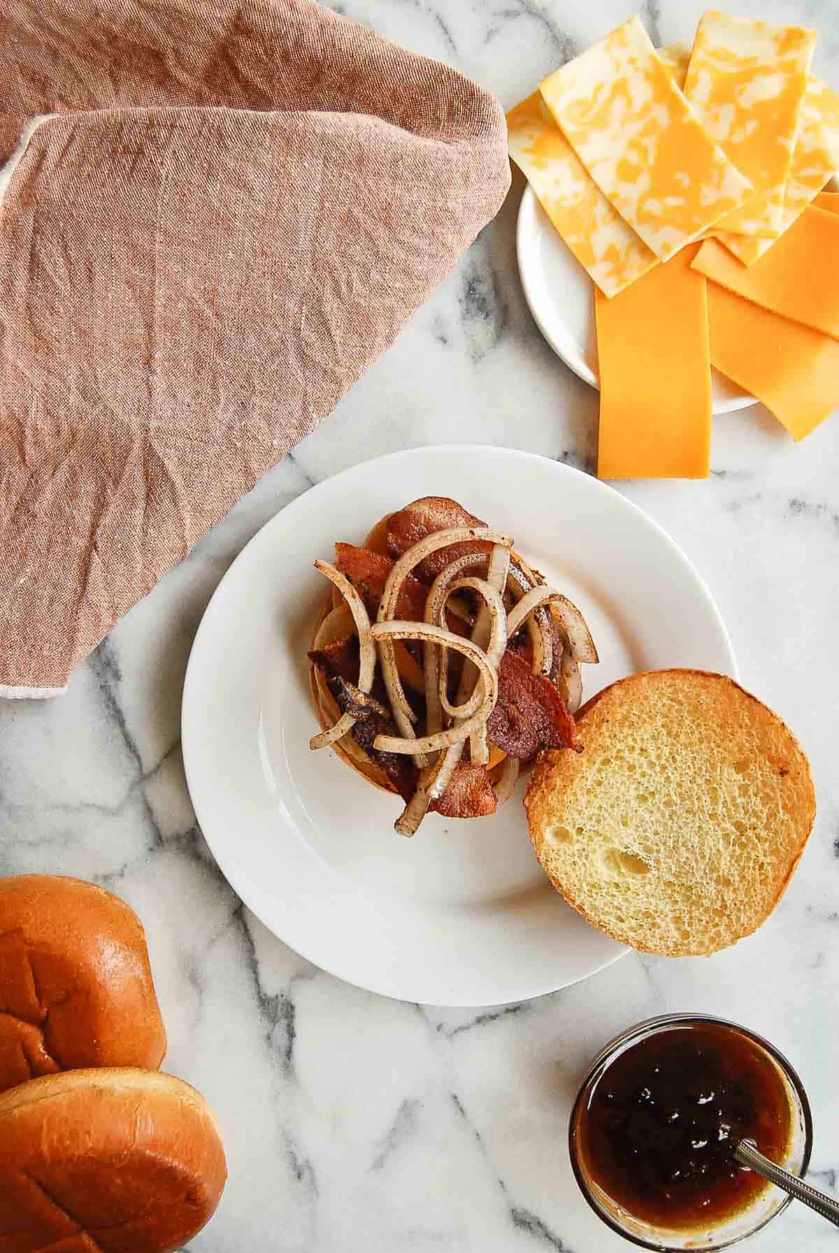 Assembly of bacon burger, with burger on bun, topped with onion and bacon.