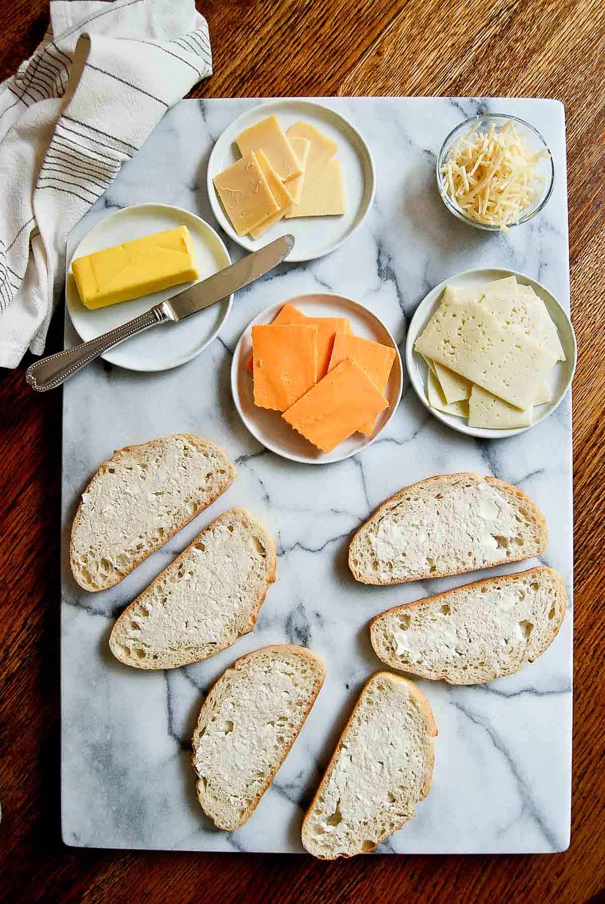 buttered bread on a marble cutting board with slices of cheese on plates.
