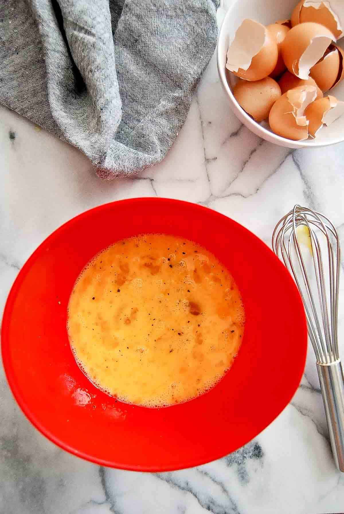 Eggs, salt and pepper in mixing bowl with whisk and bowl of cracked eggs to the side.