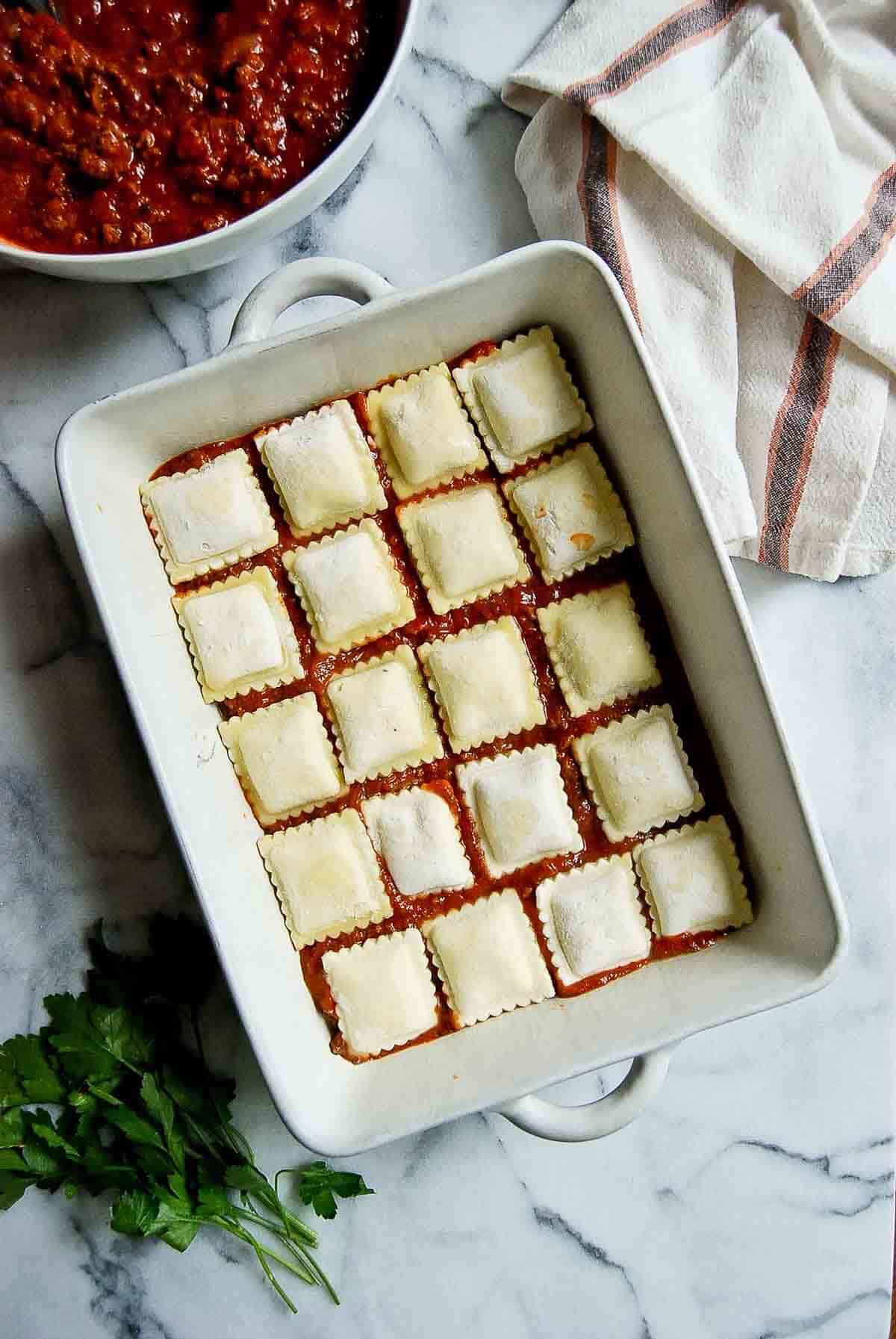 casserole dish with pasta sauce and frozen cheese ravioli being layered onto the top.