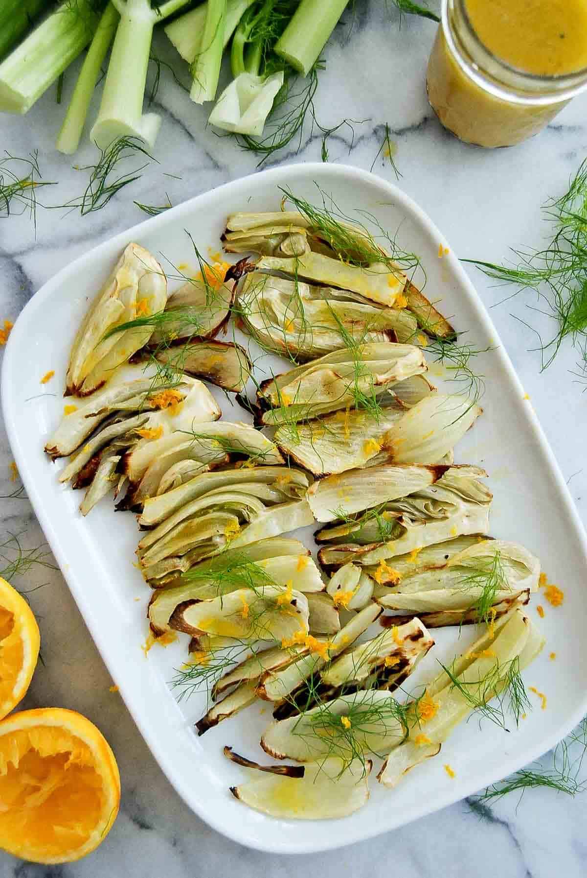 roasted fennel with orange honey dressing on serving plate with oranges, dressing and fennel stalks to the side.