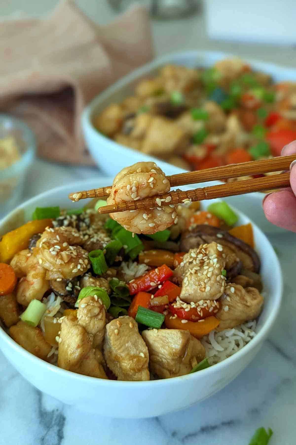 chopsticks holding piece of shrimp over bowl of chicken and shrimp stir fry with garlic ginger sauce.