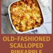 A square white dish filled with golden-brown scalloped pineapple sits on a marble surface next to a fresh pineapple and a white kitchen towel. Text below reads: Old-Fashioned Scalloped Pineapple Casserole.