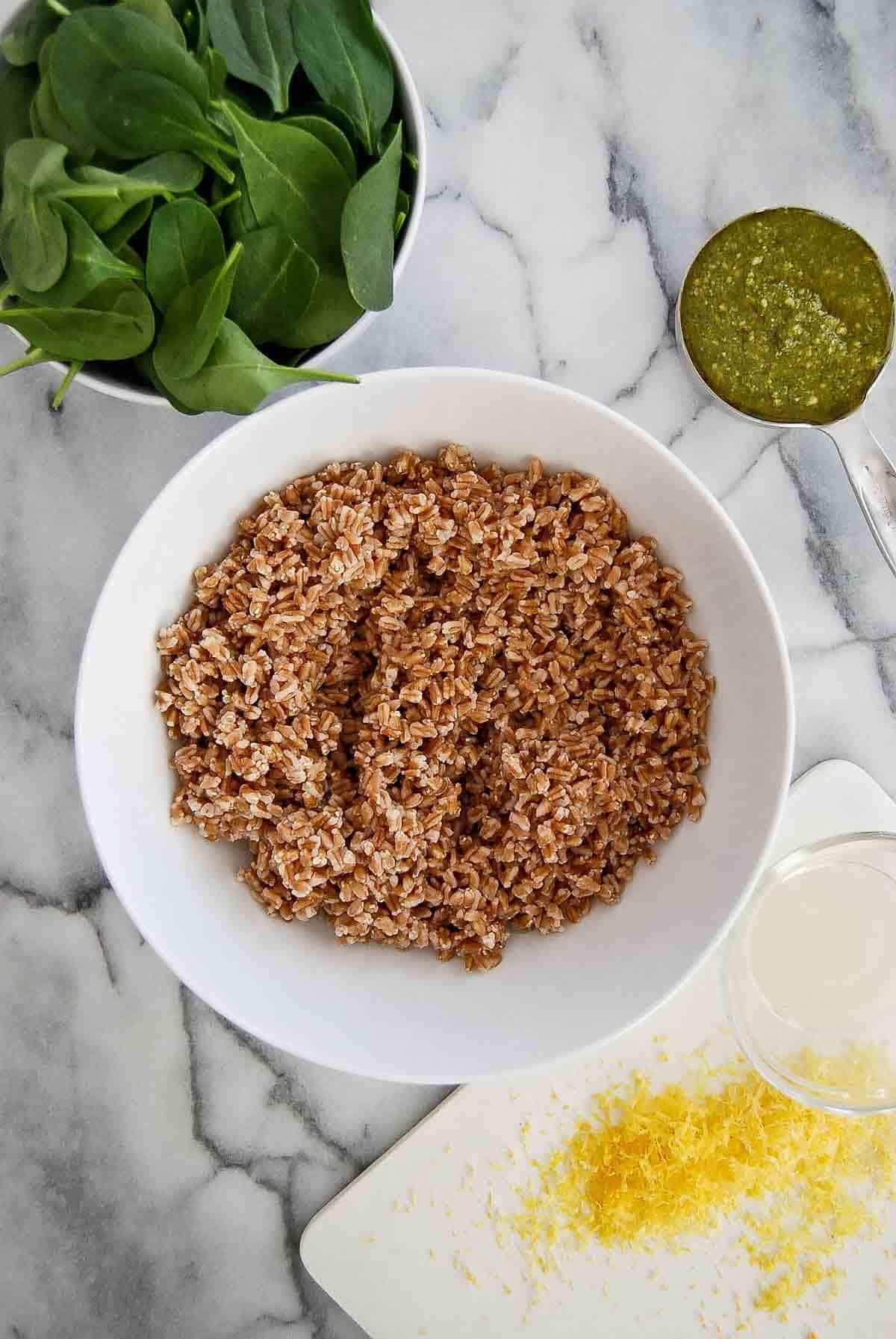 cooked farro in bowl on countertop with spinach, pesto and lemon zest on the side.