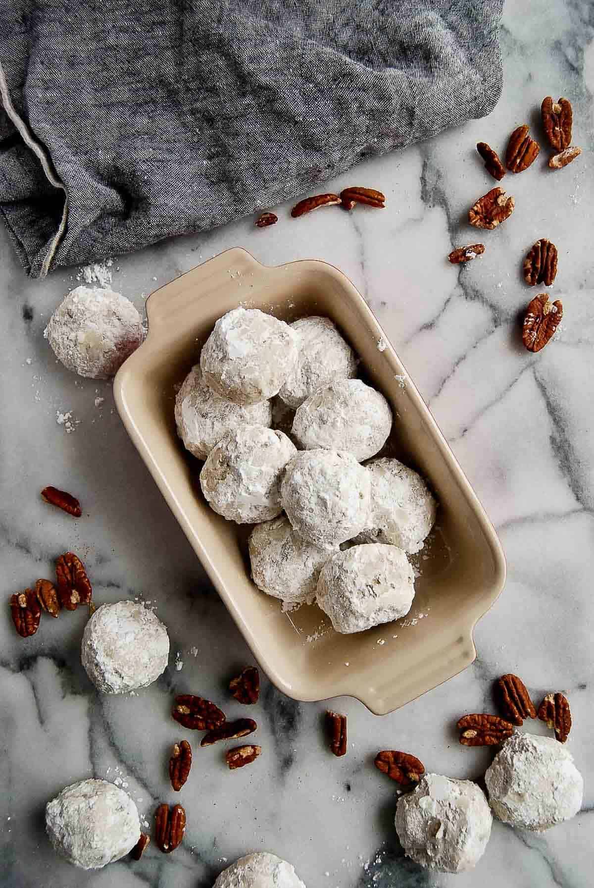 bowl of mexican wedding cookies on countertop, with more cookies and pecans scattered around.