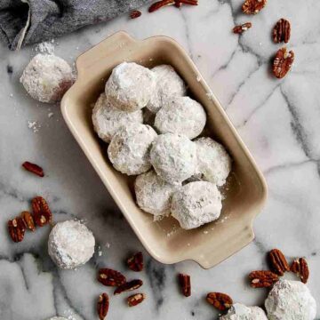 bowl of mexican wedding cookies on countertop, with more cookies and pecans scattered around.