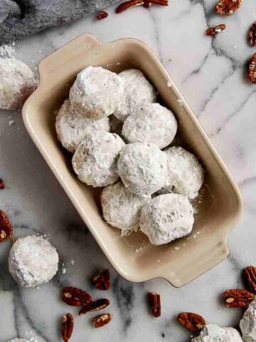 bowl of mexican wedding cookies on countertop, with more cookies and pecans scattered around.