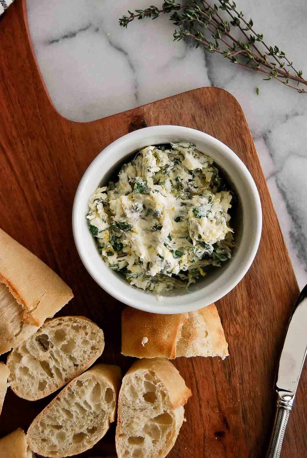 bowl of roasted garlic butter with seasoning and herbs on cutting board with bread and rosemary.