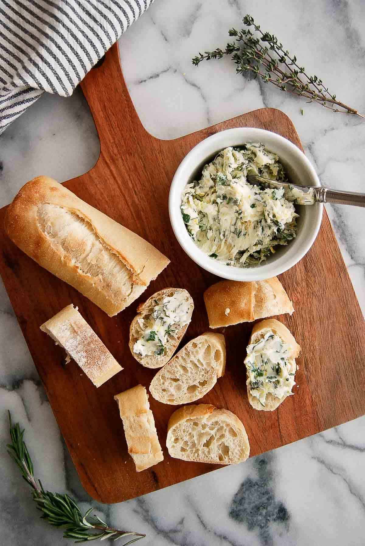 bowl of garlic butter with seasoning on cutting board with sliced baguette pieces.