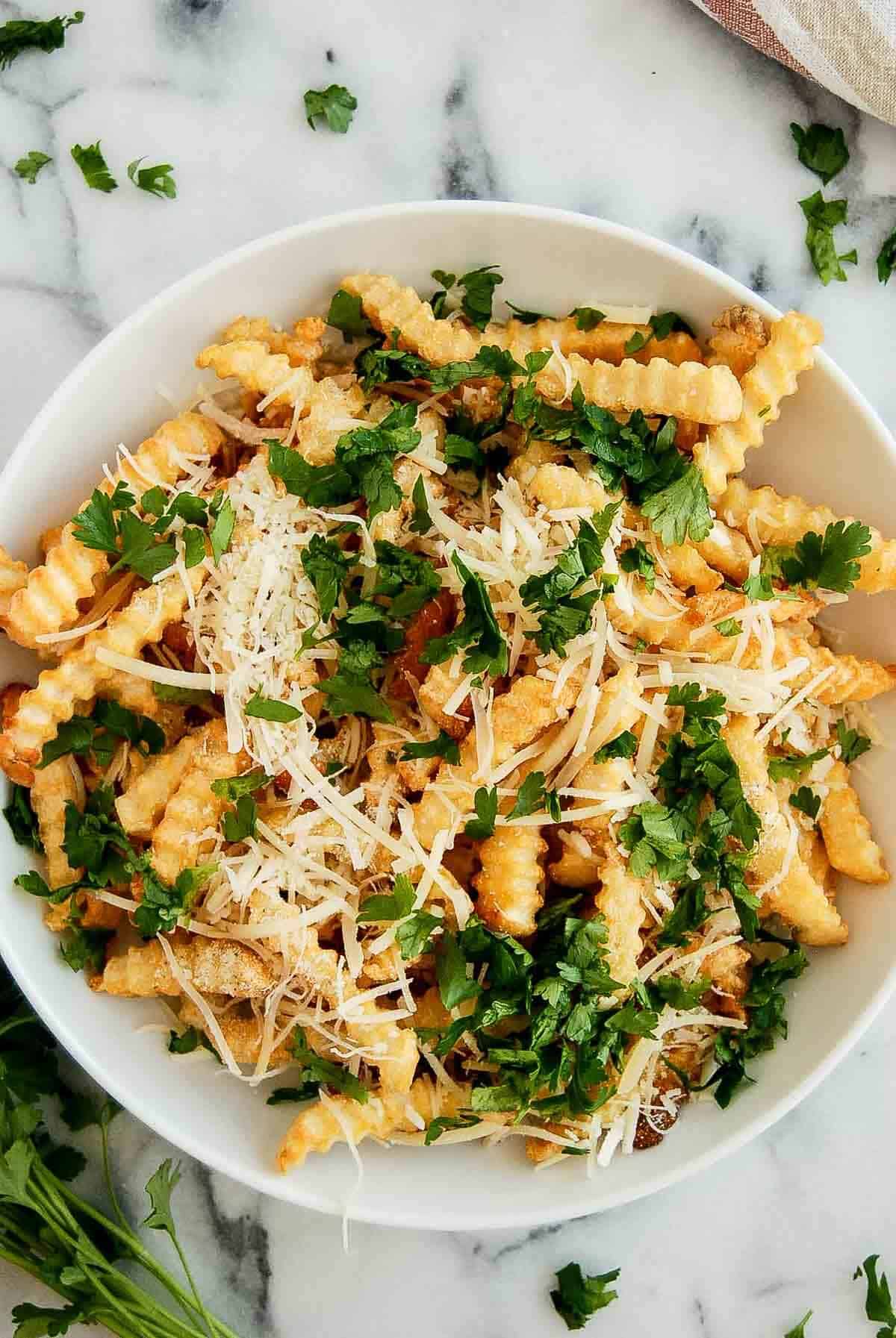 frozen french fries from air fryer with garlic salt, parmesan cheese and italian parsley in bowl on countertop.