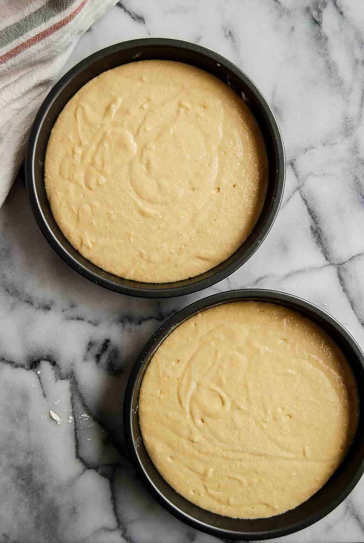 vanilla cake batter spread evenly between two cake pans on countertop.