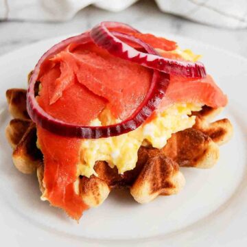 waffle breakfast sandwich with smoked salmon, cream cheese, eggs and red onion on plate.