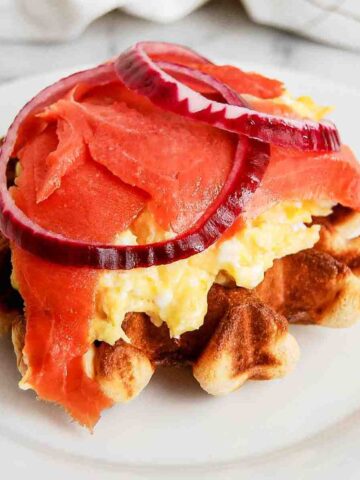 waffle breakfast sandwich with smoked salmon, cream cheese, eggs and red onion on plate.