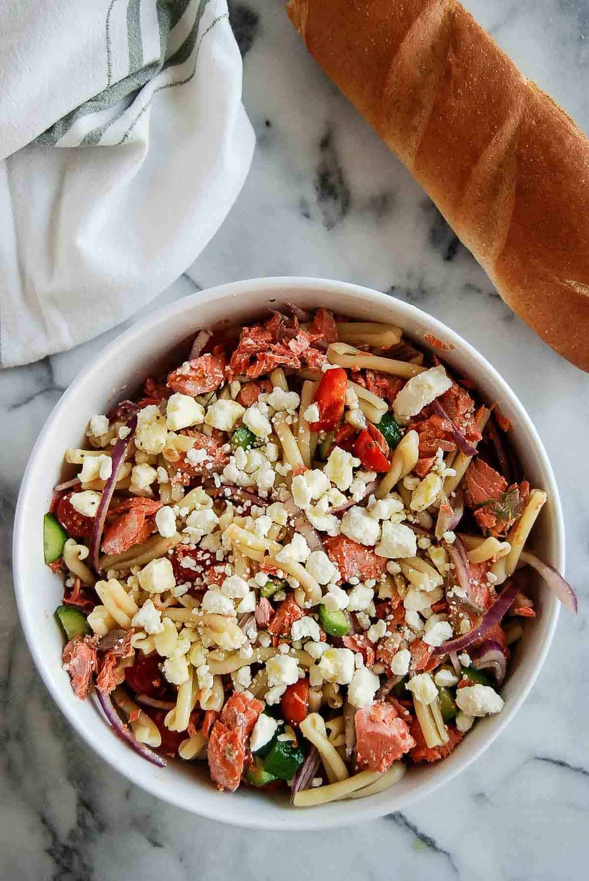 salmon pasta salad with veggies and feta in bowl on cutting board with baguette on the side.
