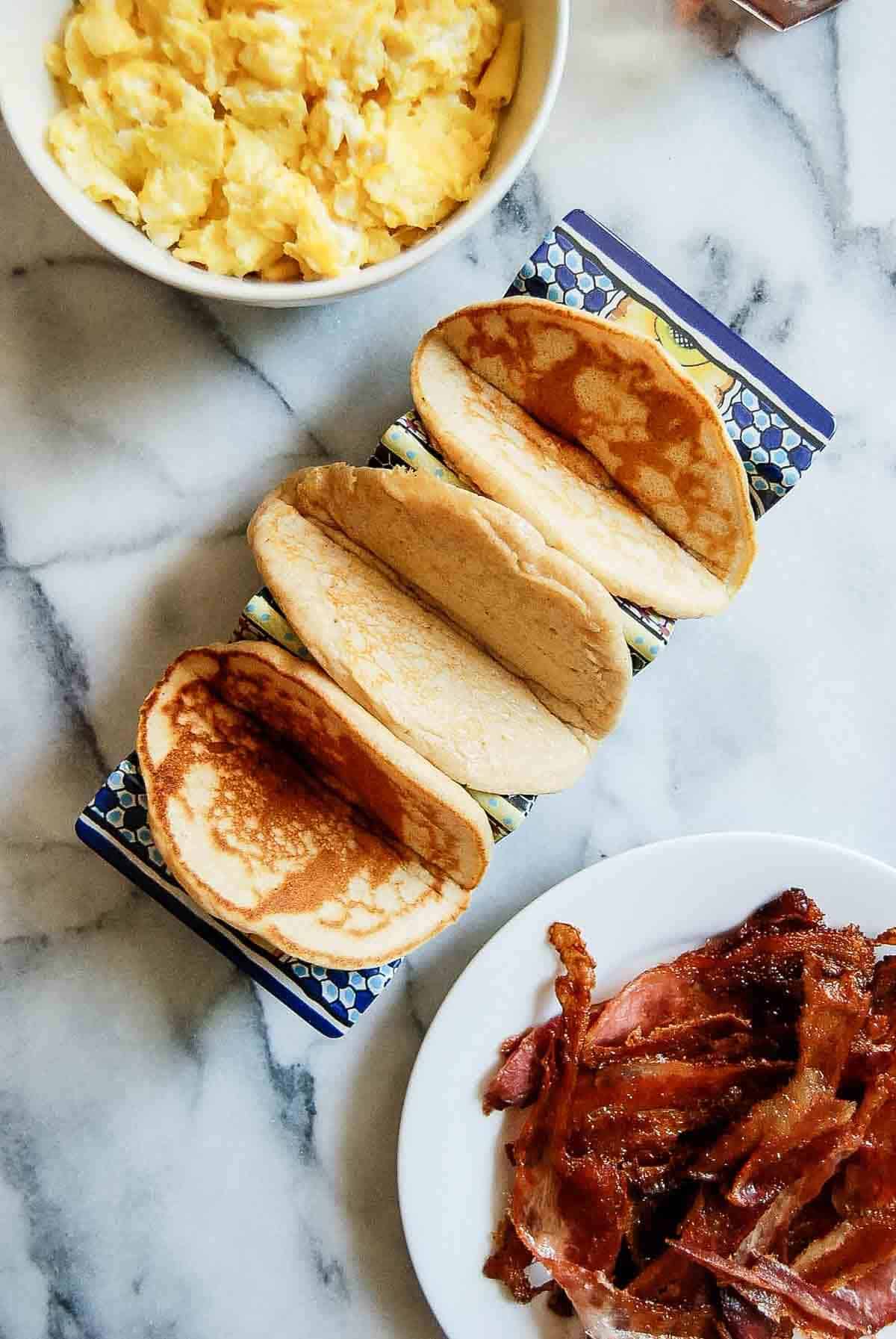 closeup of pancake taco shells with bacon and eggs on the side.