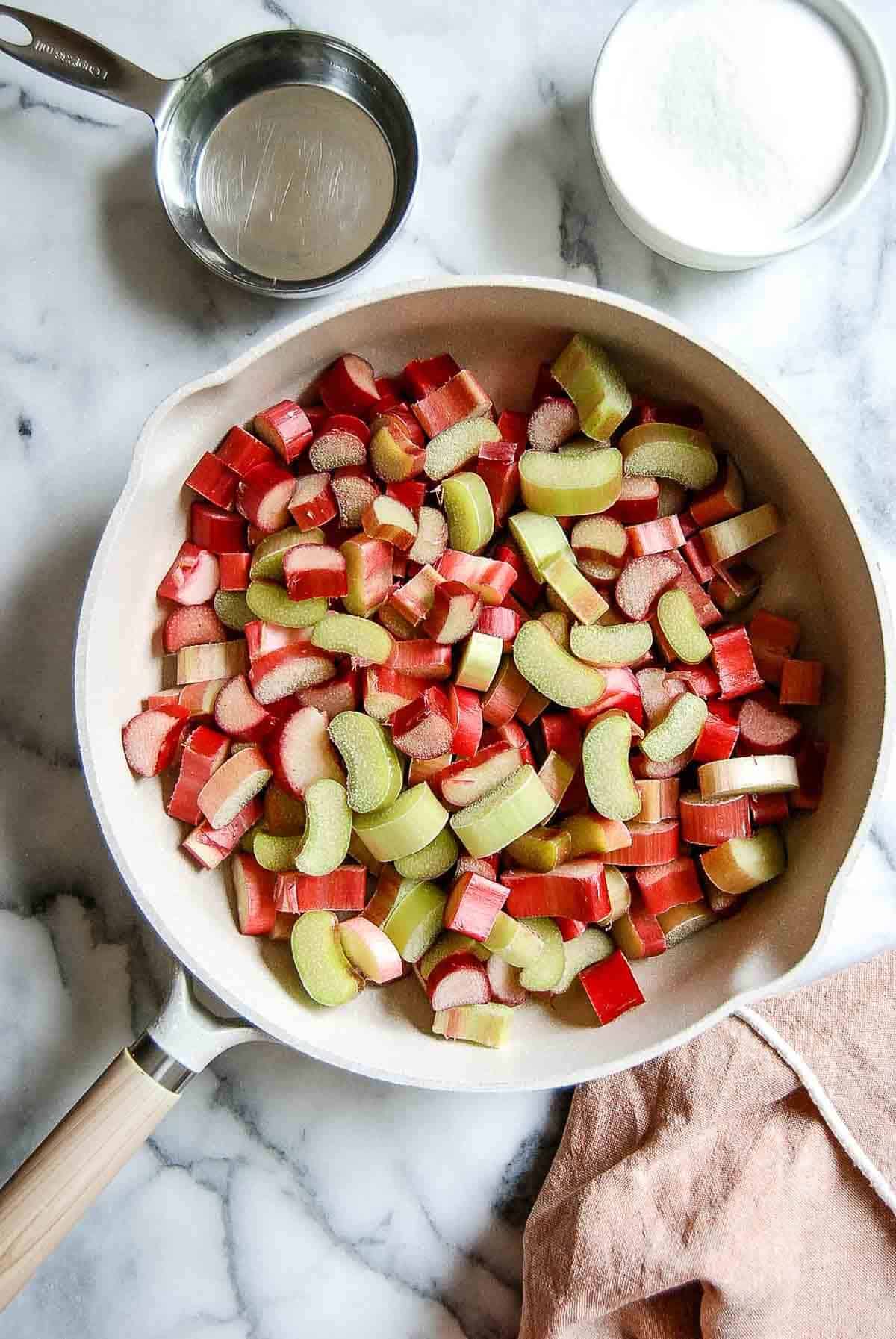 chopped rhubarb in saucepan with water and sugar on the side.