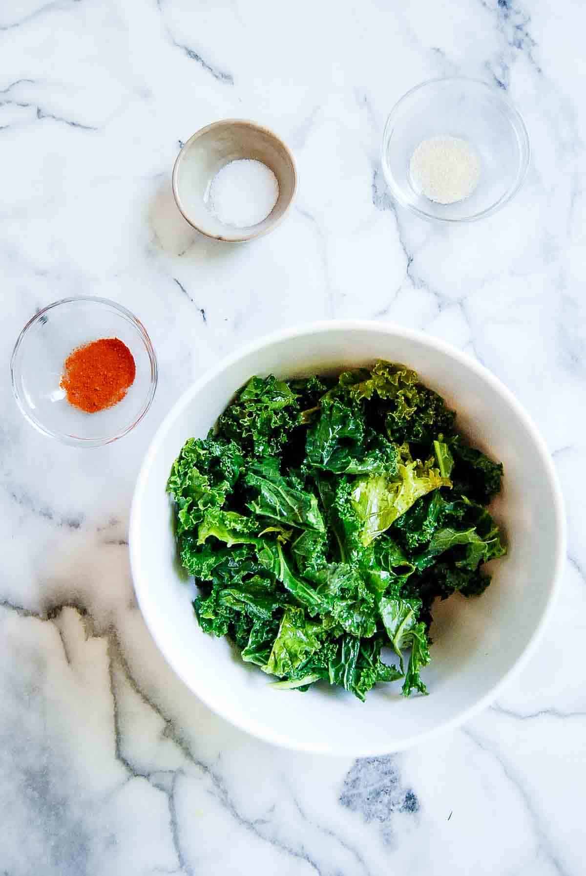 kale pieces massaged with oil in bowl, with spices on the side.