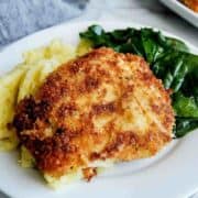 A white plate with crispy chicken fritta, mashed potatoes, and sautéed spinach sits on a marble surface with a fork nearby.