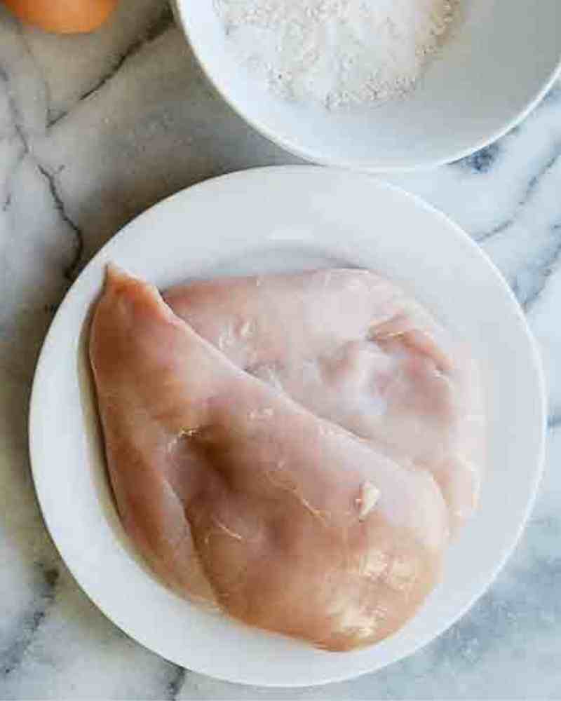 Raw chicken breasts on a white plate placed on a marble surface, with a bowl of flour visible in the upper right corner-perfect for preparing crispy chicken fritta at home.