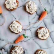 carrot cake cookies with coconut pecan cream cheese icing on countertop with decorative carrots on the sides.