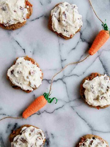 carrot cake cookies with coconut pecan cream cheese icing on countertop with decorative carrots on the sides.