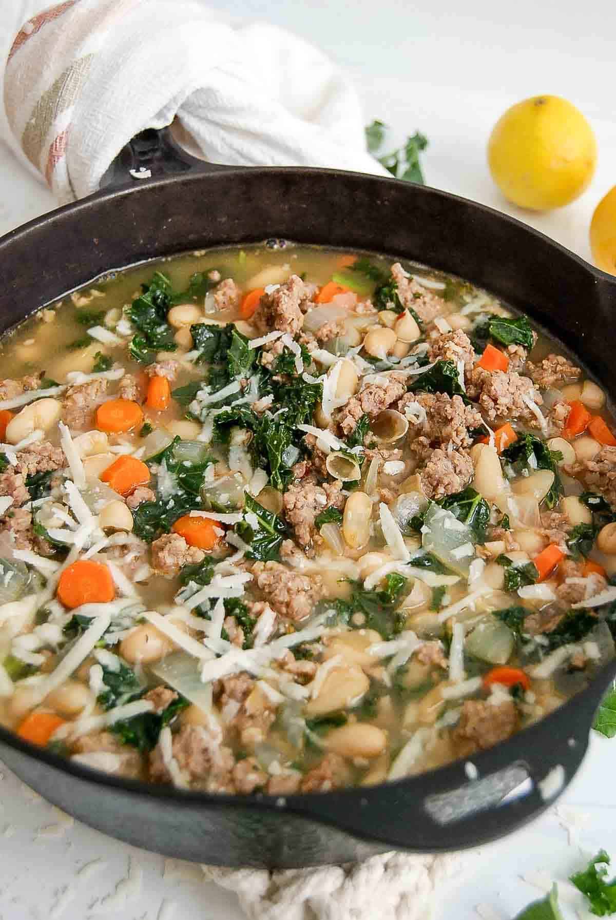 side view of pot of sausage, lake and white bean soup with shredded parmesan on the top and lemons on the side.